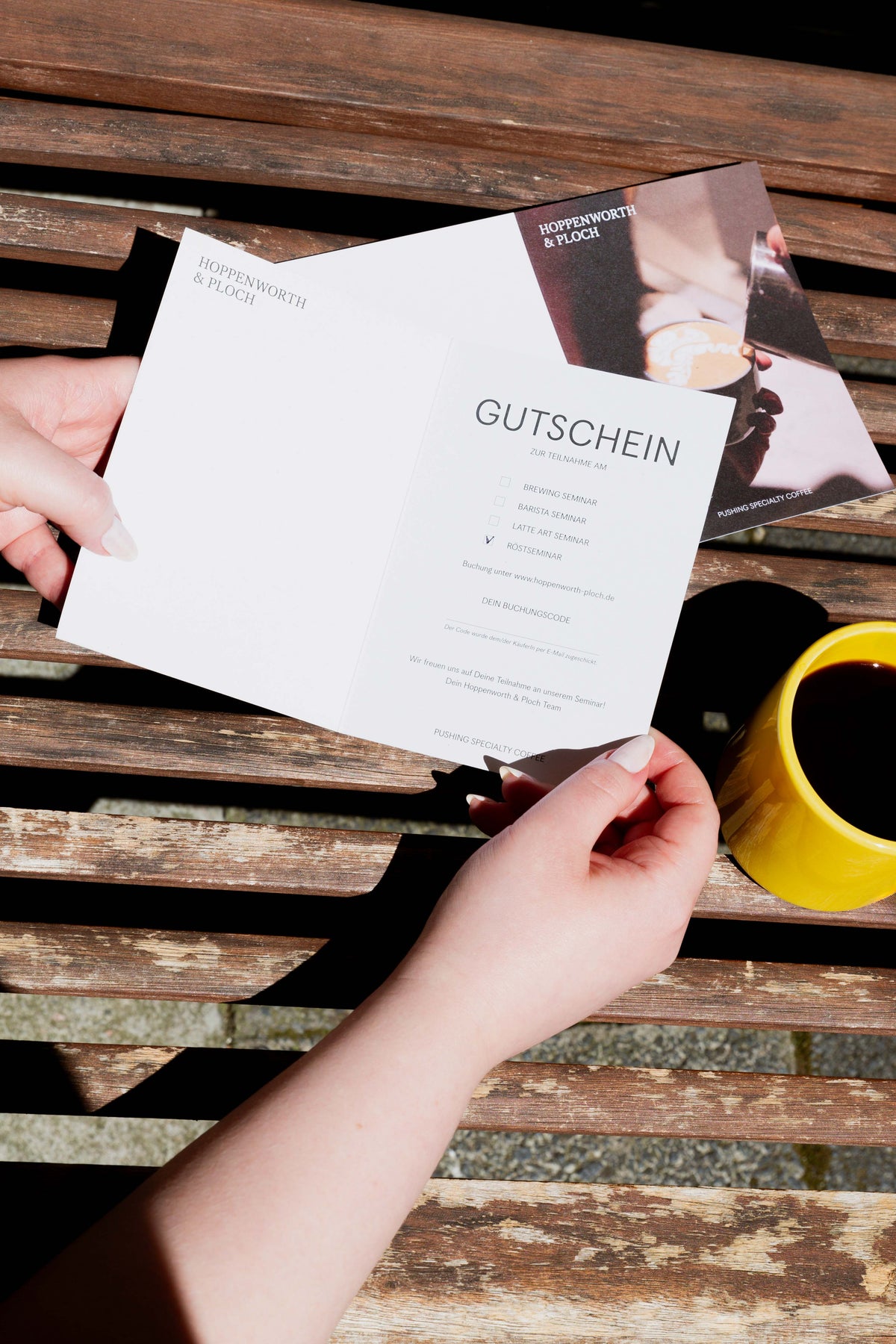 Eine Person hält eine Gutscheinkarte, im Hintergrund liegt sie von der anderen Seite, und ein gelber Kaffeepott ist sichtbar.