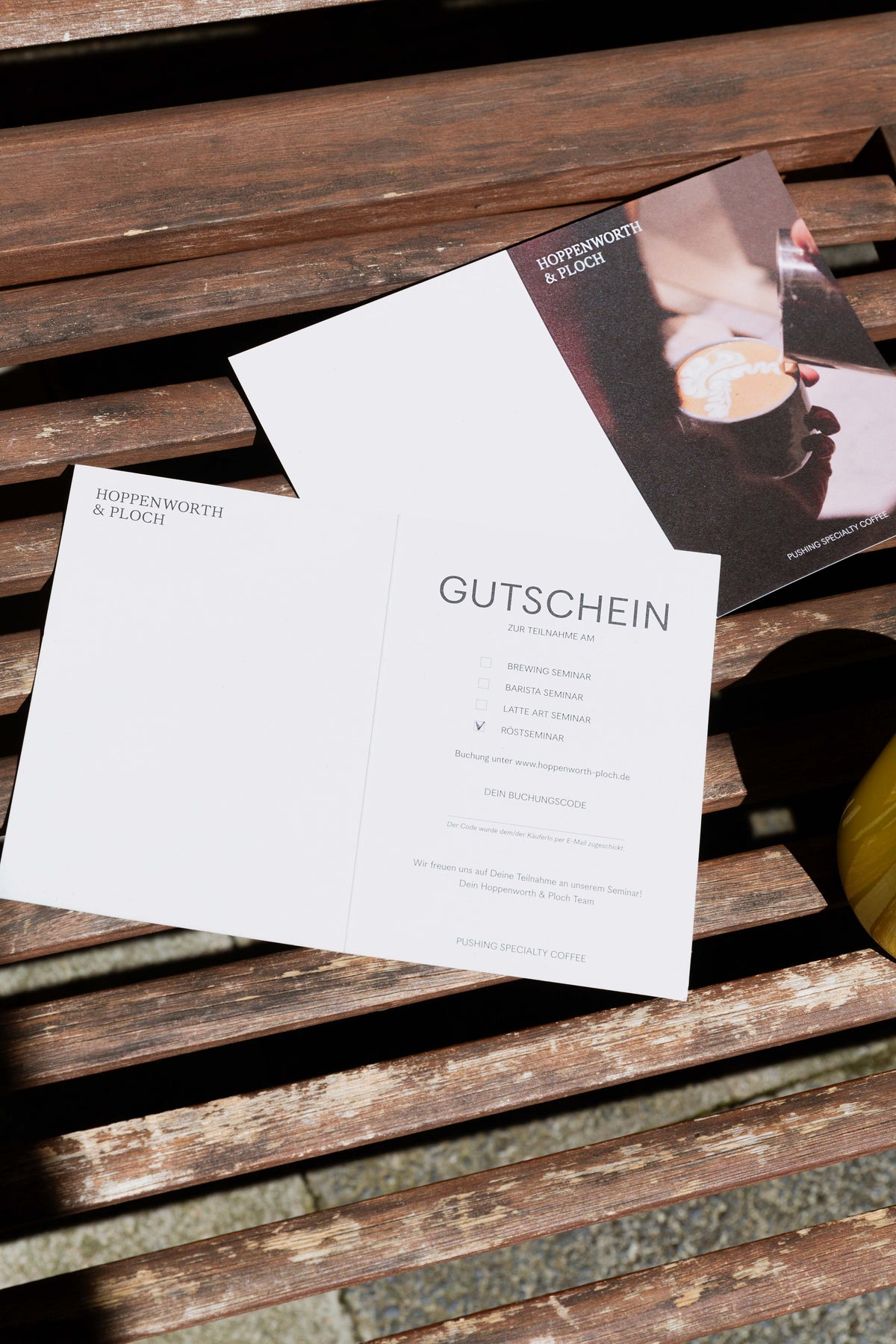 eine Gutscheinkarte, im Hintergrund liegt sie von der anderen Seite, und ein gelber Kaffeepott ist sichtbar.